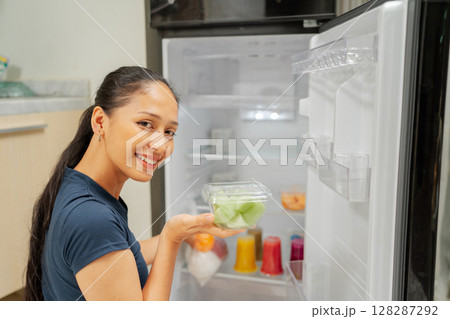 A young woman skillfully organizes fresh produce in her stylish refrigerator A young woman skillfully organizes fresh produce in her stylish refrigerator 128287292