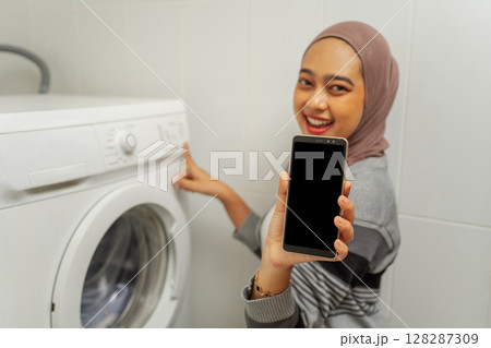 A Happy User is Showcasing Their Smartphone Alongside a Washing Machine at Home A Happy User is Showcasing Their Smartphone Alongside a Washing Machine at Home 128287309