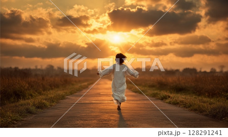 Joyful boy in white garment runs along picturesque road in golden sunset, surrounded by lush fields, embodying innocence 128291241