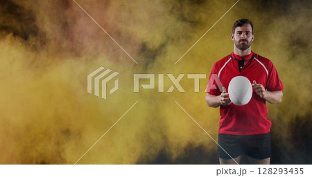 Rugby player in red jersey holding ball, standing confidently against colorful smoke 128293435