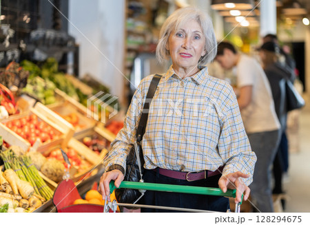 In food store, senior woman with trolley looking for something on shelf of showcase In food store, senior woman with trolley looking for something on shelf of showcase 128294675