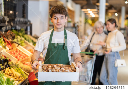 Guy employee in dark green apron stands in sales area with box of mushrooms 128294813