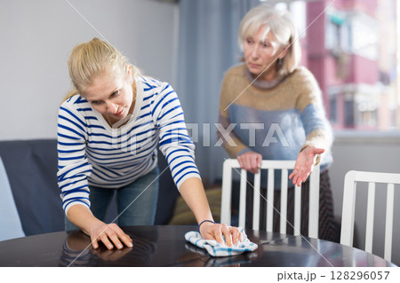Elderly mother is unhappy with the way her adult daughter dusts the table in room Elderly mother is unhappy with the way her adult daughter dusts the table in room 128296057