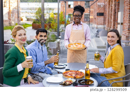 A smiling waitress serves pizza to a group of friends at an outdoor cafe. A smiling waitress serves pizza to a group of friends at an outdoor cafe. 128299509