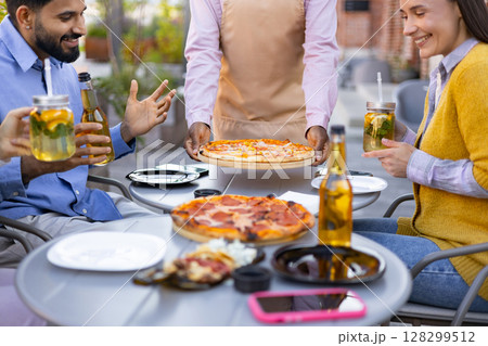 A diverse group of people enjoying pizza, drinks, and each other's company outdoors. A waiter is serving pizza. 128299512