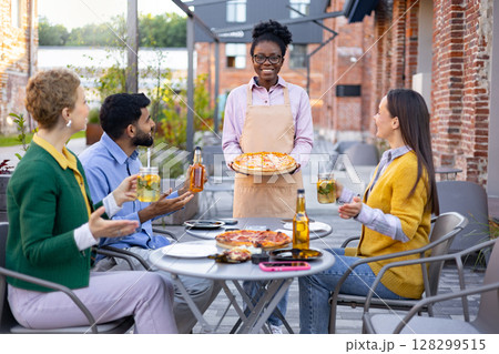 A smiling waitress serves pizza to a group of friends at an outdoor cafe, enjoying drinks and the sunny day. 128299515