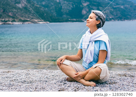 Mature woman meditating near ocean, enjoying tranquility and fresh air on mountain beach Mature woman meditating near ocean, enjoying tranquility and fresh air on mountain beach 128302740