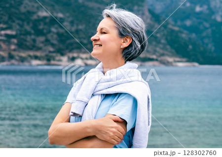 Happy Senior Woman Smiling Outdoors Near the Sea with Crossed Arms and Wind in Her Hair 128302760