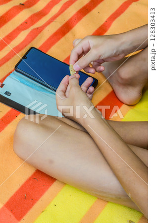 Child sits on striped blanket holding snack chip and smartphone during summer leisure. High quality photo Child sits on striped blanket holding snack chip and smartphone during summer leisure. High quality photo 128303413