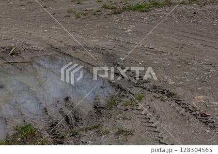 Tire tracks in muddy ground near a small puddle reflecting surrounding vegetation during a sunny day in early spring Tire tracks in muddy ground near a small puddle reflecting surrounding vegetation during a sunny day in early spring 128304565