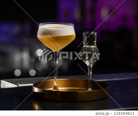 A beautifully presented golden-hued cocktail with a rich foam top is served alongside a small shot glass of clear liquid, all resting on a chic golden tray A beautifully presented golden-hued cocktail with a rich foam top is served alongside a small shot glass of clear liquid, all resting on a chic golden tray 128304621