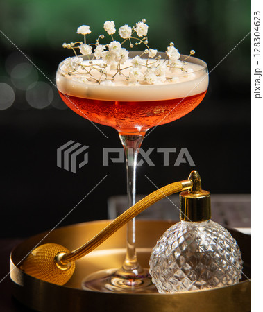 blush-toned cocktail with a frothy top is elegantly garnished with delicate white baby's breath flowers, presented on a golden tray alongside a vintage-style perfume atomizer 128304623