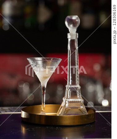A classic martini in a frosted glass stands beside a unique Eiffel Tower-shaped decanter, both presented on a golden tray 128306169