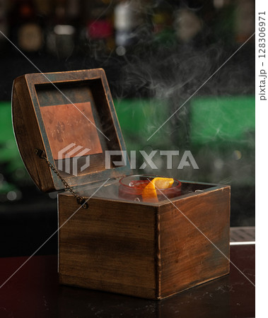A smoky, dark red alcoholic cocktail with ice and an orange peel garnish is presented in a rocks glass nestled inside an open wooden box. 128306971