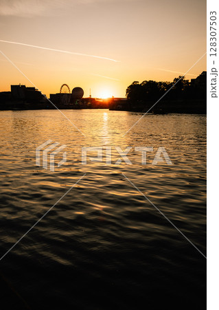 Vertical screen: shadowed riverside view with distant silhouette of Ferris wheel and urban buildings reflecting on water surface, capturing moody atmosphere and gentle evening light. Summer City Mood 128307503
