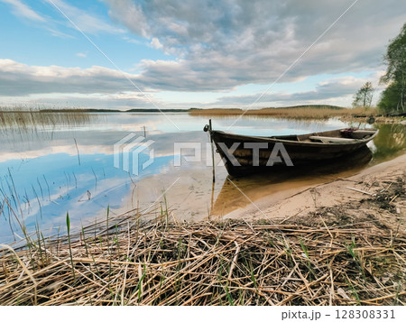 Old wooden fishing boat is moored on shore of calm lake, reflecting blue and cloudy sky, surrounded by reeds and dry grass, creating peaceful and evocative scene. Wooden fishing boat rests on lakeside 128308331