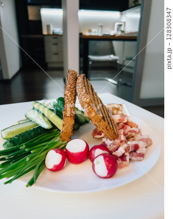 Toasted rye bread, bacon, radish, green onions and cucumber slices arranged on white plate, creating simple and appetizing meal in modern kitchen setting. Crispy bread, bacon, radishes, green onions Toasted rye bread, bacon, radish, green onions and cucumber slices arranged on white plate, creating simple and appetizing meal in modern kitchen setting. Crispy bread, bacon, radishes, green onions 128308347