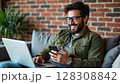 A smiling man wearing glasses relaxes on a sofa, simultaneously using a laptop and a smartphone with a brick wall background. - Generic AI Image 128308842