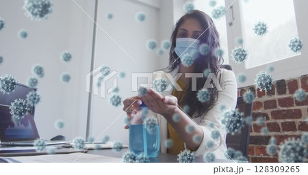 Image of covid 19 cells floating over biracial woman wearing face mask disinfecting her hands in off 128309265