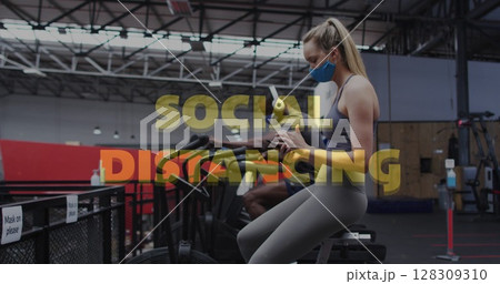 Social distancing text against caucasian woman wearing face mask using smartphone at gym 128309310