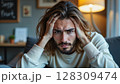 Portrait of a frustrated young man holding his head with both hands, sitting indoors on a couch  Generic AI Image 128309474