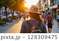 Back view of a woman wearing a hat and backpack walking on a sunny street with people in the background - Generic AI Image 128309475
