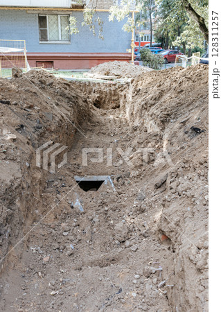 A trench dug in front of the facade of an apartment building. Excavation work. Repair work on a heating main. 128311257