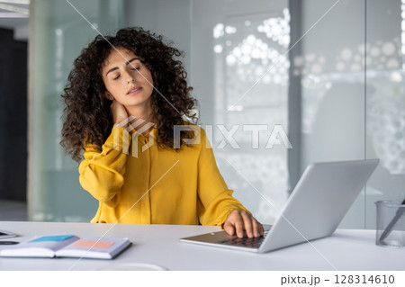 Businesswoman feeling neck pain sitting at desk using laptop. Expression shows workplace discomfort. Scene represents stress and strain in office settings. Businesswoman feeling neck pain sitting at desk using laptop. Expression shows workplace discomfort. Scene represents stress and strain in office settings. 128314610