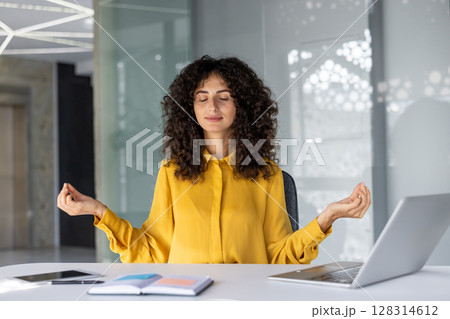 Young woman meditating at office desk with closed eyes. Practicing mindfulness and relaxation for stress relief during work. Laptop, notebook nearby, promoting calm and focus in busy environment. 128314612