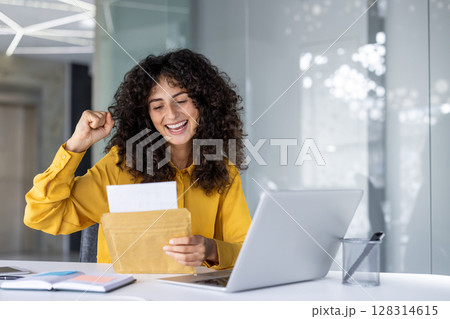 Happy woman in yellow shirt rejoicing positive news at desk with laptop. She holds envelope with excited expression, symbolizing career achievement and satisfaction at work. Modern workspace ambiance 128314615
