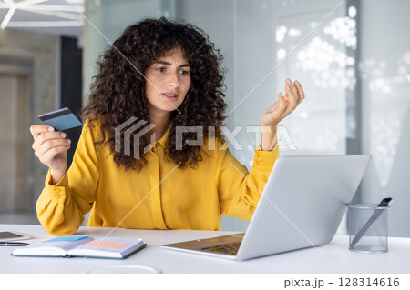 Woman sitting at desk holding credit card while looking at laptop with frustrated expression. Indicates online payment issue or possible fraud, confusion over transaction or account error. 128314616