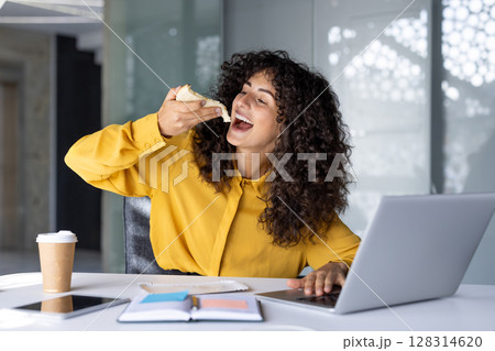 Confident Hispanic businesswoman takes snack break while working at desk with laptop and coffee. Shows balance between work and personal time in modern office environment. Confident Hispanic businesswoman takes snack break while working at desk with laptop and coffee. Shows balance between work and personal time in modern office environment. 128314620