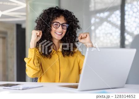 A smiling woman with curly hair wearing glasses celebrates a success while using a laptop computer. 128314652