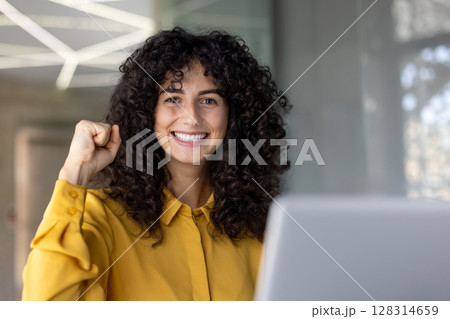 A cheerful woman with curly hair smiles with a fist raised, exuding success and confidence. She wears a yellow shirt. A cheerful woman with curly hair smiles with a fist raised, exuding success and confidence. She wears a yellow shirt. 128314659