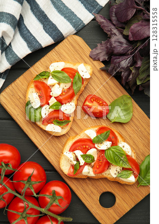Tasty sandwich with mozzarella cheese, tomatoes and basil on cutting board on black wooden background. Caprese Tasty sandwich with mozzarella cheese, tomatoes and basil on cutting board on black wooden background. Caprese 128315558