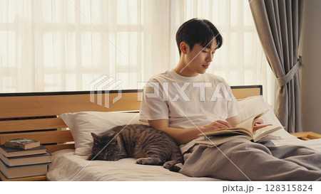 猫が寝ているベッドで本を読んでいる男性 128315824