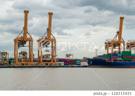 Shipping cranes work in a busy port, lifting cargo containers against a cloudy sky. A vital hub of trade and transport. 128315935