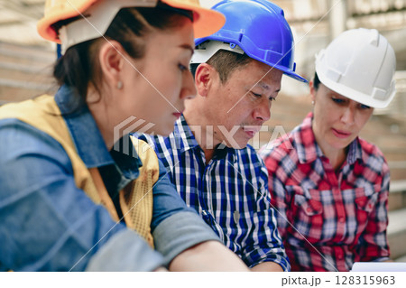 Three construction workers collaborating on project plans at a job site with hard hats and focused expressions. 128315963