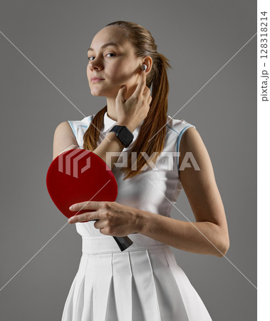 Focused female table tennis player adjusting earbud while holding paddle on gray background Focused female table tennis player adjusting earbud while holding paddle on gray background 128318214