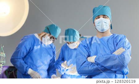 Surgeon in blue scrubs and mask standing in operating room with medical team working in background. 128320119