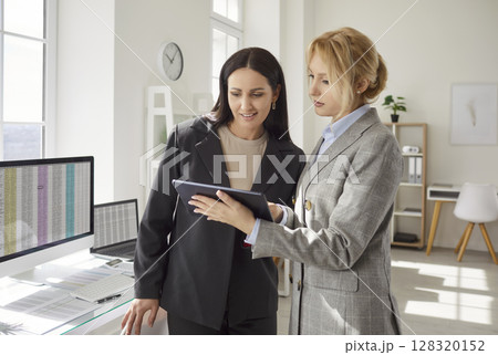 Two businesswomen standing in office with tablet, discussing financial report and analyzing data. Two businesswomen standing in office with tablet, discussing financial report and analyzing data. 128320152