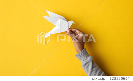 Hand holds paper dove against bright yellow background, representing creativity, peace, childhood and origami art. 128320694