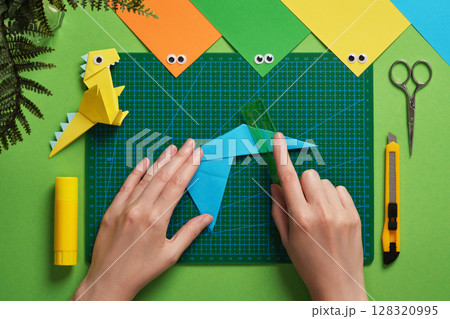 Person folds blue origami paper on a green cutting mat using a ruler. Nearby are colorful paper, googly eyes, a yellow paper dinosaur, scissors, a craft knife, glue stick 128320995