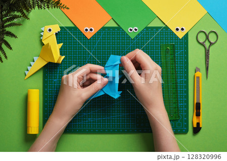 Hands fold blue origami dinosaur on a green cutting mat. Nearby are colorful paper, googly eyes, a yellow paper dinosaur, scissors, a craft knife, glue stick Hands fold blue origami dinosaur on a green cutting mat. Nearby are colorful paper, googly eyes, a yellow paper dinosaur, scissors, a craft knife, glue stick 128320996