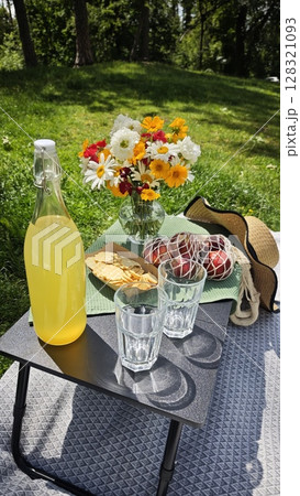 Bottle of lemonade, glasses, crackers and fresh flowers on picnic table in sunny green park 128321093