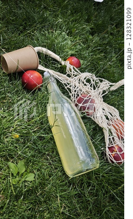 Bottle of lemonade, paper cup and peaches in eco mesh bag on green grass in sunny park 128321099