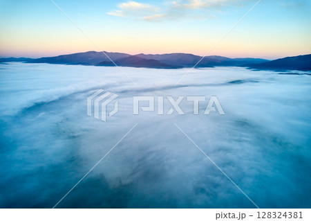 Breathtaking aerial view at dawn, showcasing vast sea of clouds enveloping mountain landscape. Subtle hues of rising sun paint sky in soft pastels, creating tranquil and ethereal scene. 128324381