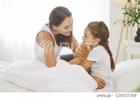 Cute little girl waking up in morning holding teddy bear tightly and talking sincerely to her mother Cute little girl waking up in morning holding teddy bear tightly and talking sincerely to her mother 128327169