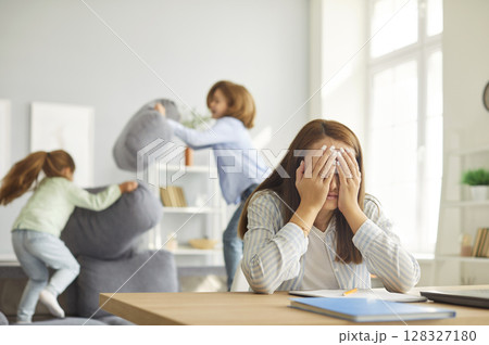 Exhausted mother sitting at desk with head in hands as kids jumping and playing loudly on sofa. 128327180