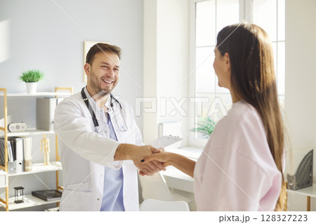 Smiling male doctor in white medical uniform meets to consult female patient, hand shaking, male GP  128327223